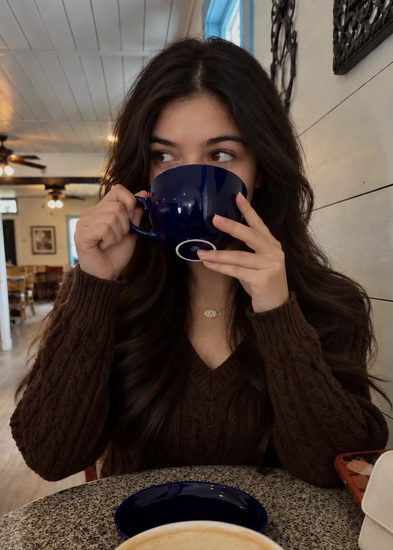 Suhani in a brown cable-knit sweater, sipping from a navy cup at a café table, a latte in front of her